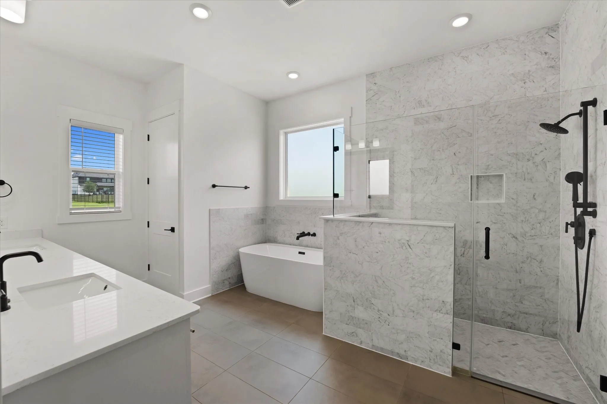 Spacious bathroom featuring a frameless glass walk-in shower with matte black fixtures, a freestanding soaking tub, dual vanity with undermount sinks and quartz countertops, and large format tile flooring