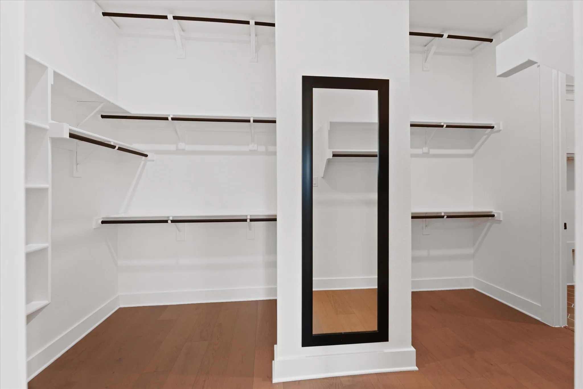 Spacious walk-in closet featuring wood-finish flooring, white built-in shelving, multiple hanging rods, and a full-length mirror