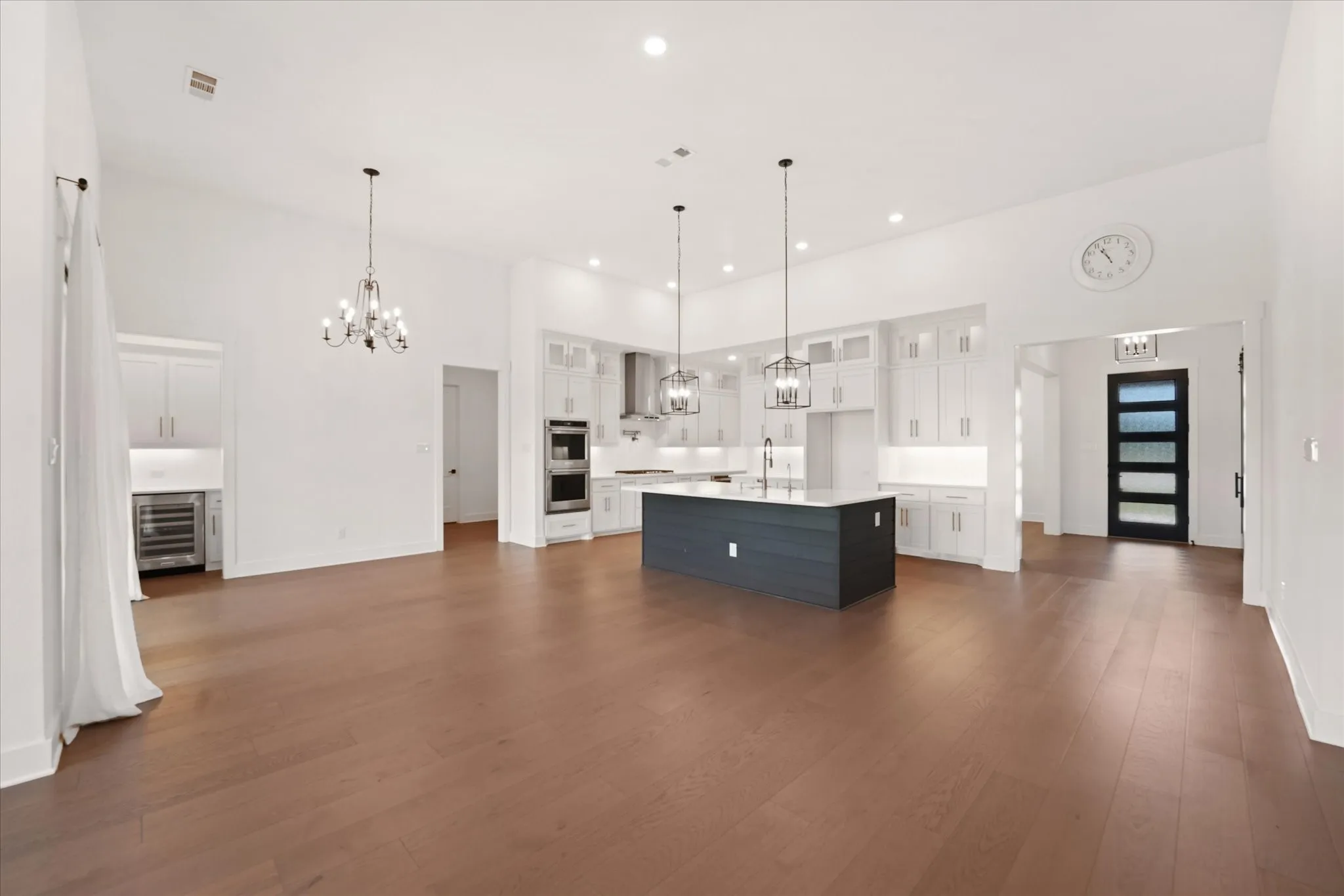 Spacious interior featuring wood-finish flooring, a contemporary kitchen island with pendant lighting, white cabinetry, integrated double ovens, and a beverage refrigerator