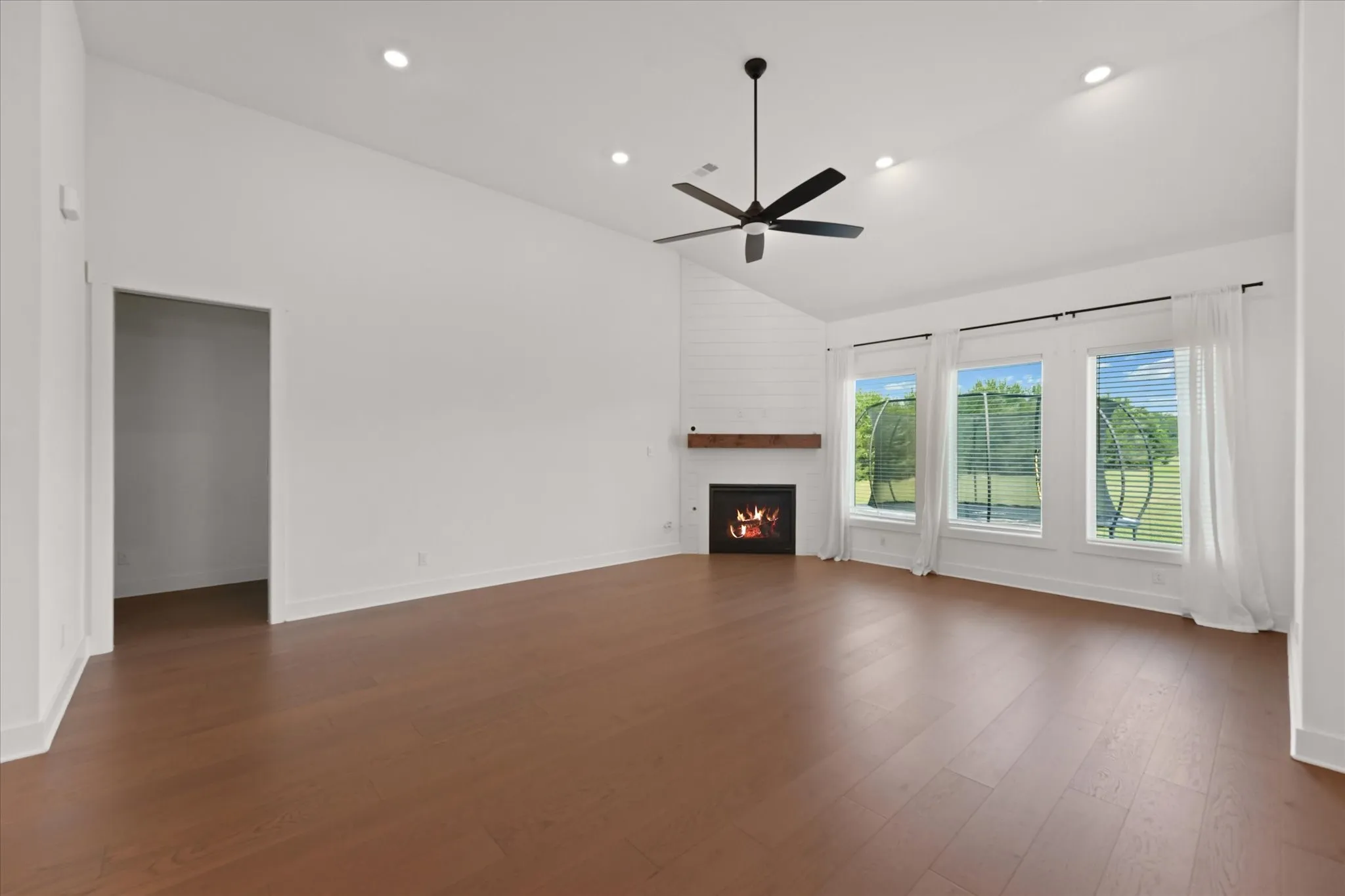 Spacious room featuring rich wood-finish flooring, a vaulted ceiling with recessed lighting and ceiling fan, and multiple windows with blinds and sheer curtains