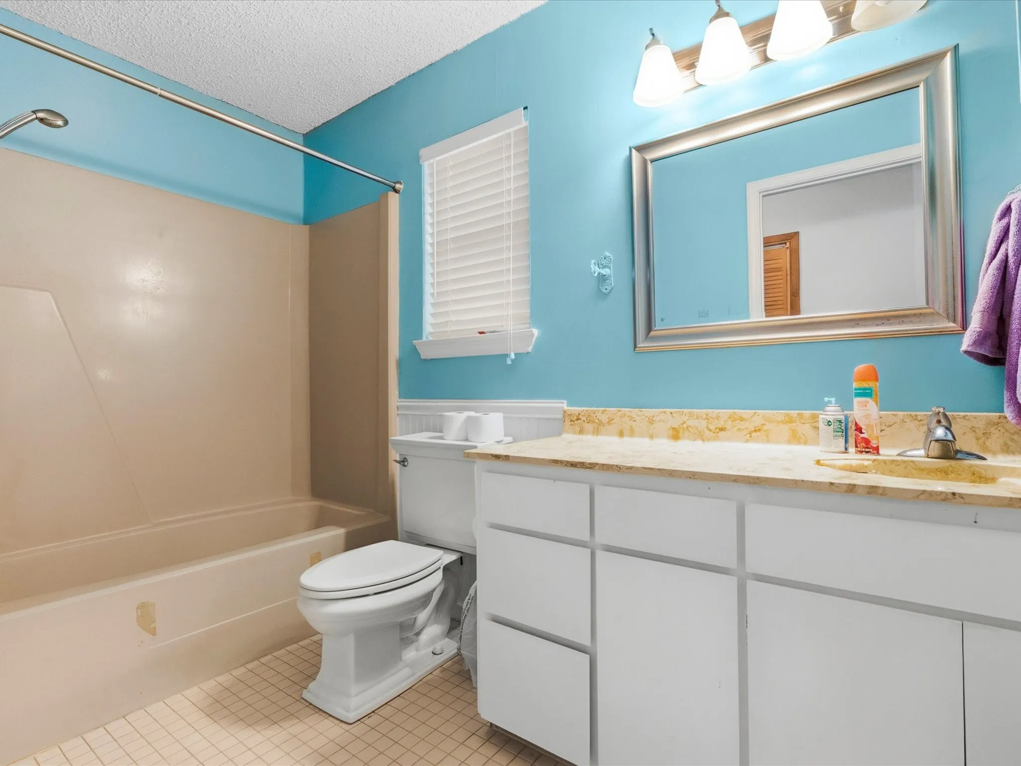 Bathroom featuring a cream-colored tub/shower combination, a vanity with white cabinetry, a light-colored stone countertop, a rectangular mirror with a silver-toned frame, and tile flooring