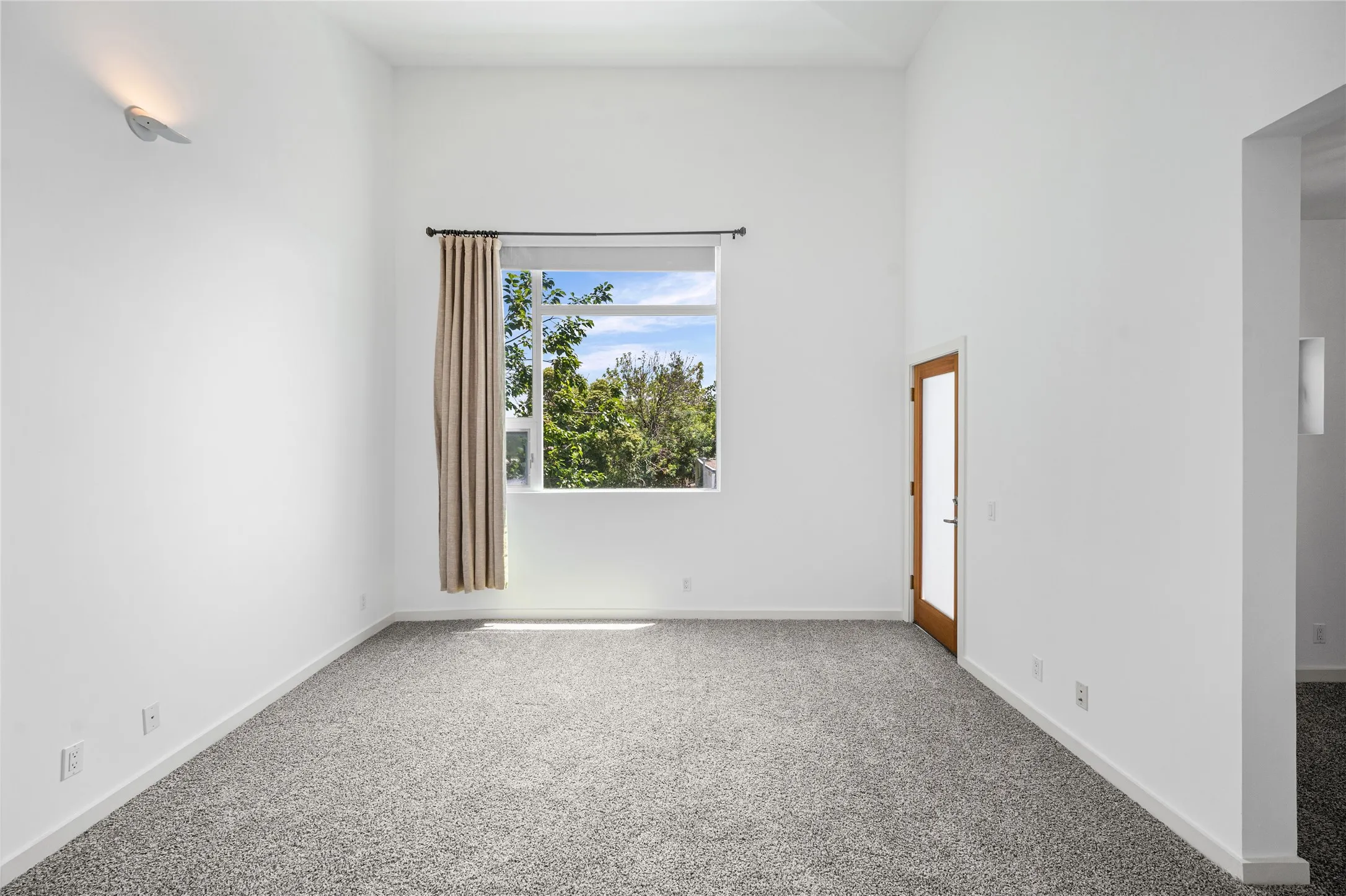Spacious interior featuring high ceilings, light gray carpeting, a large window, a partial-pane wood-finish door, and a modern wall sconce