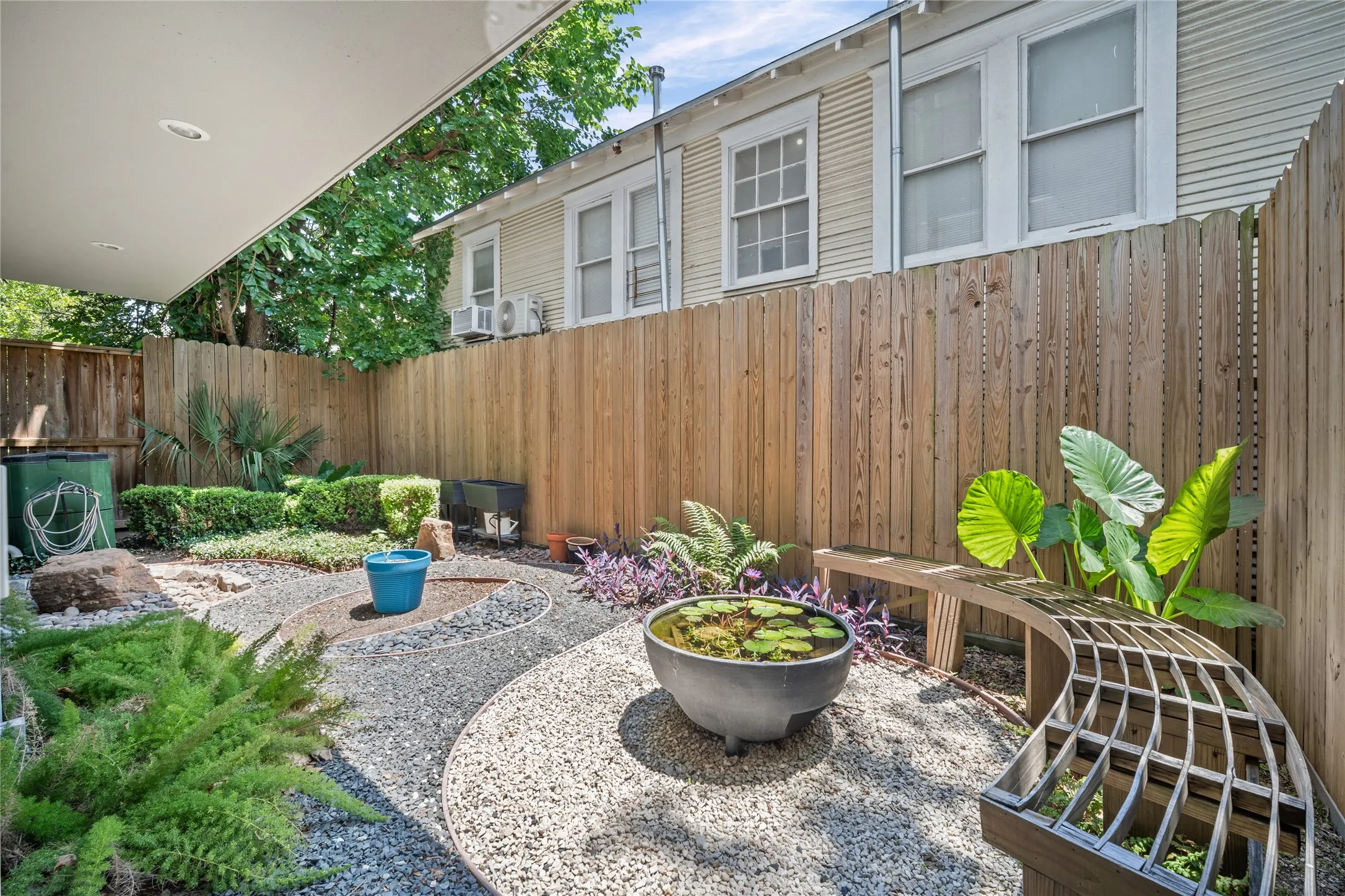 Private backyard oasis featuring diverse plantings, gravel pathways, a water feature, and a curved slatted bench