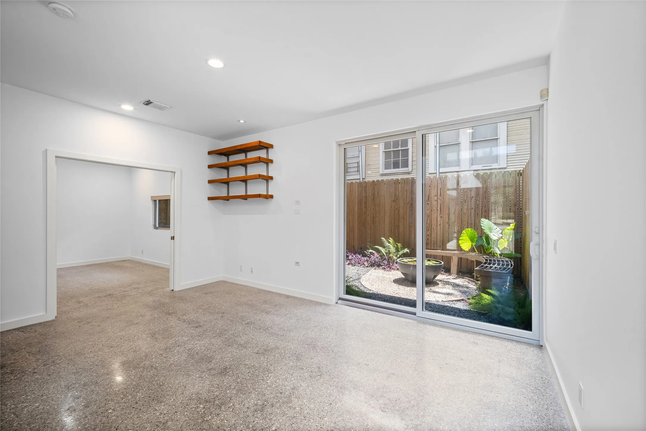 Spacious room featuring polished concrete flooring, recessed lighting, built-in floating wood shelves, and a sliding glass door leading to a private outdoor gravel patio with lush plantings and a wooden bench