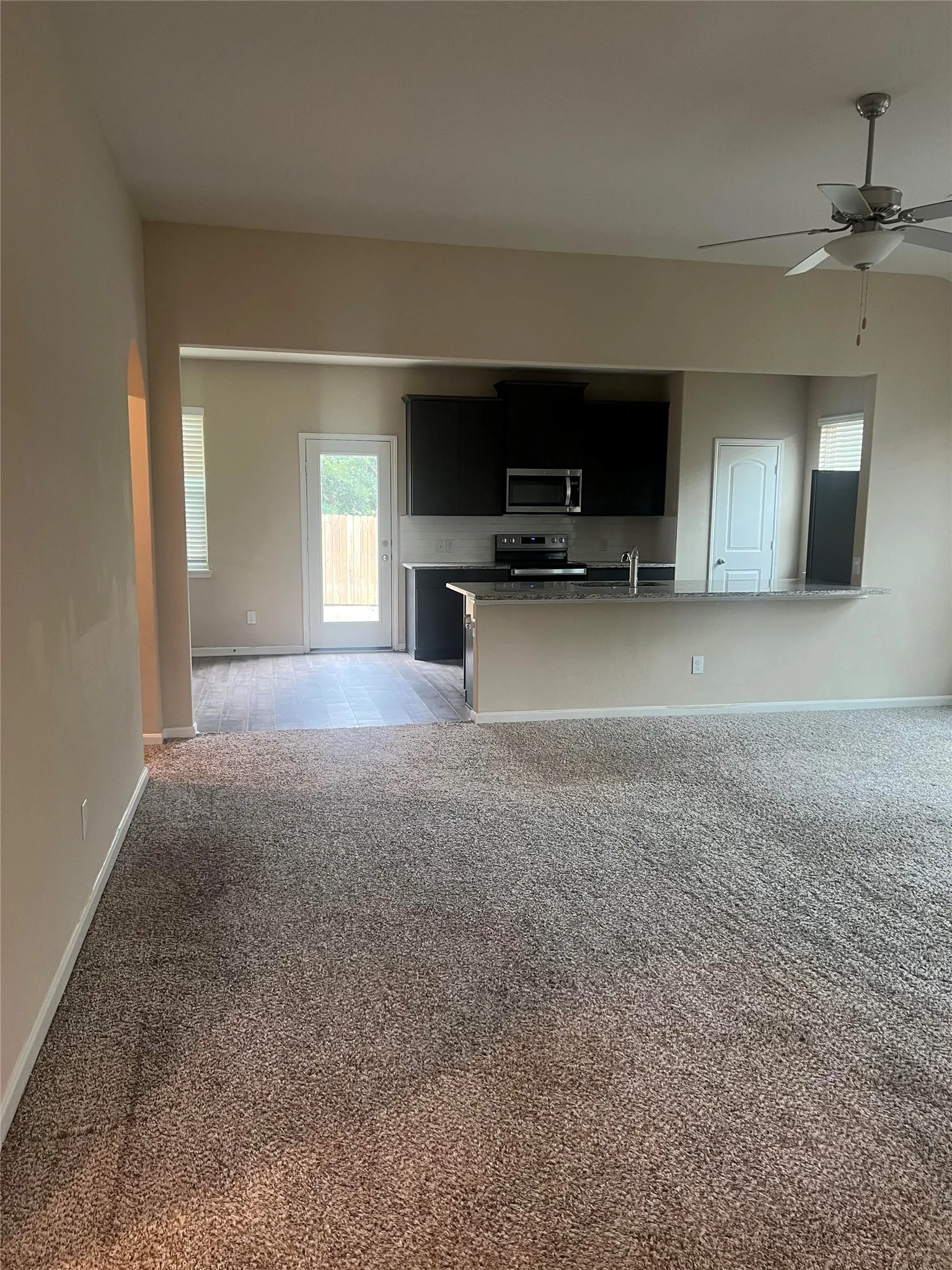 Open-concept living area featuring plush carpeting, a ceiling fan with integrated lighting, and a view into the kitchen