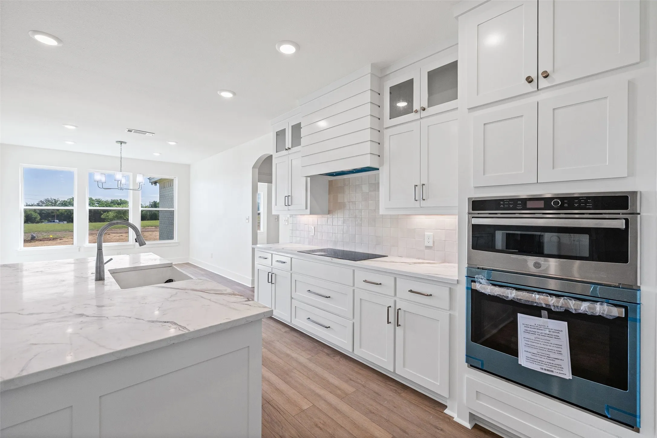 Contemporary kitchen featuring bright white cabinetry, a large center island with an undermount sink, built-in stainless steel appliances, wood-finish flooring, and a tile backsplash