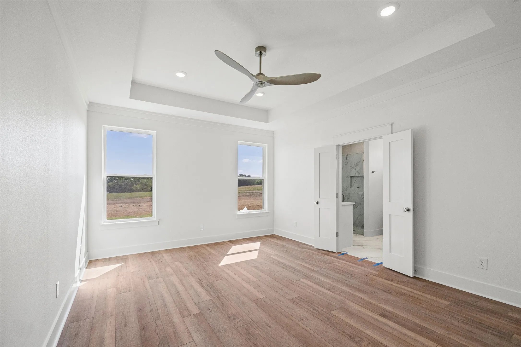 Spacious room featuring wood-finish flooring, a tray ceiling with recessed lighting and a ceiling fan, and two windows providing natural illumination