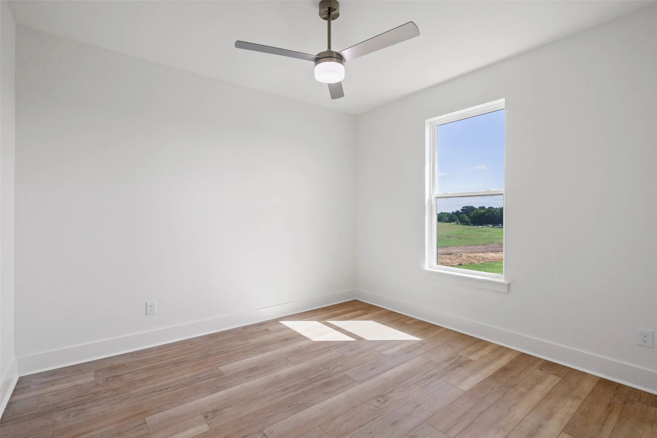 Room featuring wood-finish flooring, a contemporary ceiling fan with integrated lighting, and a window framing a pastoral landscape
