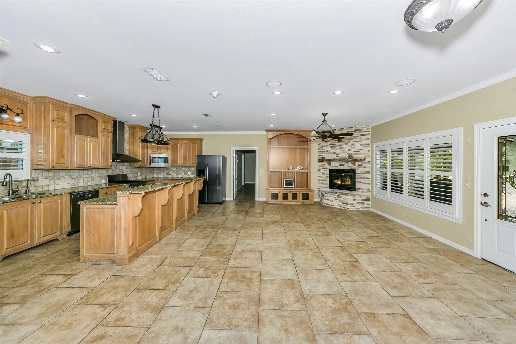 Expansive open-concept room featuring a kitchen with wood cabinetry and an island with bar seating, a stone-clad fireplace, and tile flooring