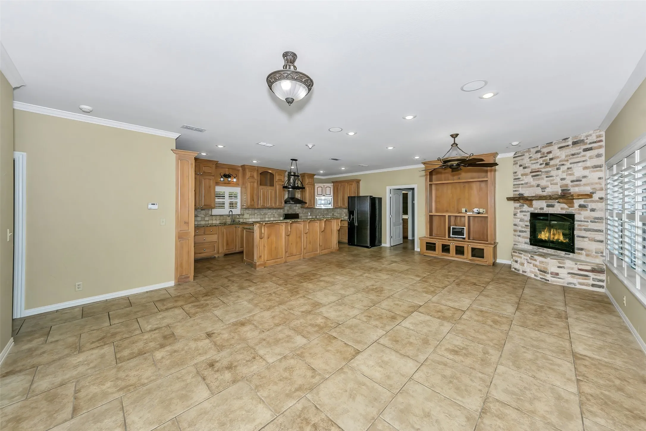 Expansive open-concept living area featuring large format tile flooring, a built-in media center with ceiling fan, and a stone-clad fireplace