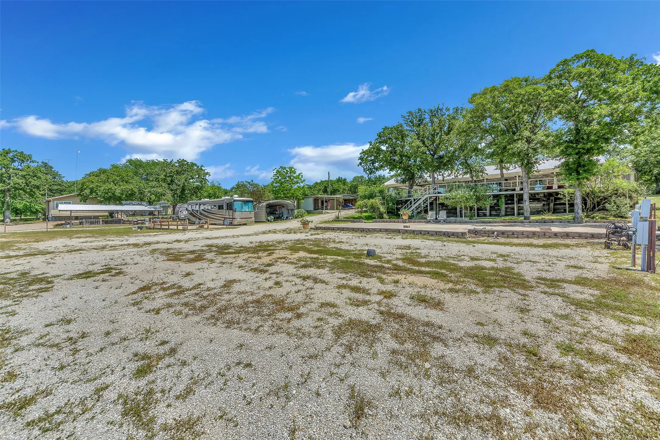 Expansive gravel lot with mature trees, a multi-level deck, and a long covered structure