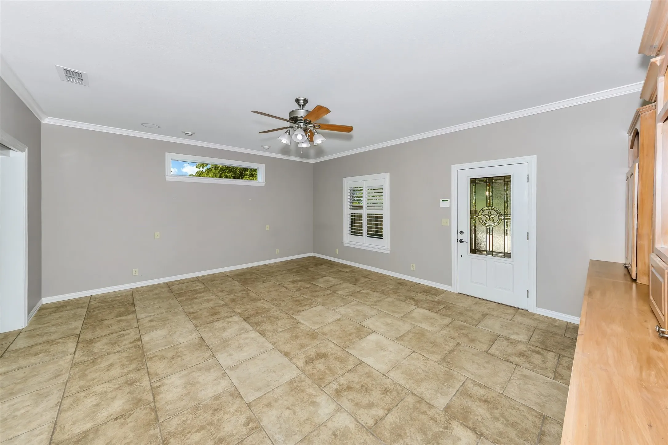 Spacious room featuring neutral wall paint, tile flooring, crown molding, a decorative front entry door with leaded glass, and a ceiling fan