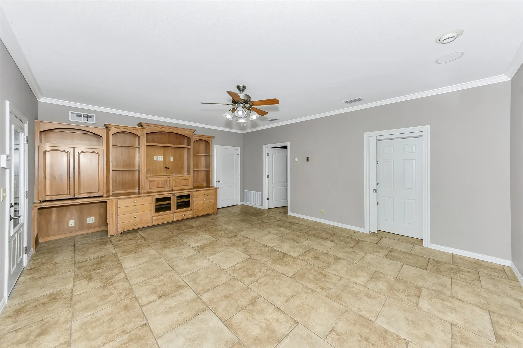 Spacious room featuring tile flooring, light gray walls, a built-in entertainment center with extensive cabinetry and shelving, a ceiling fan with integrated lighting, and crisp white crown molding