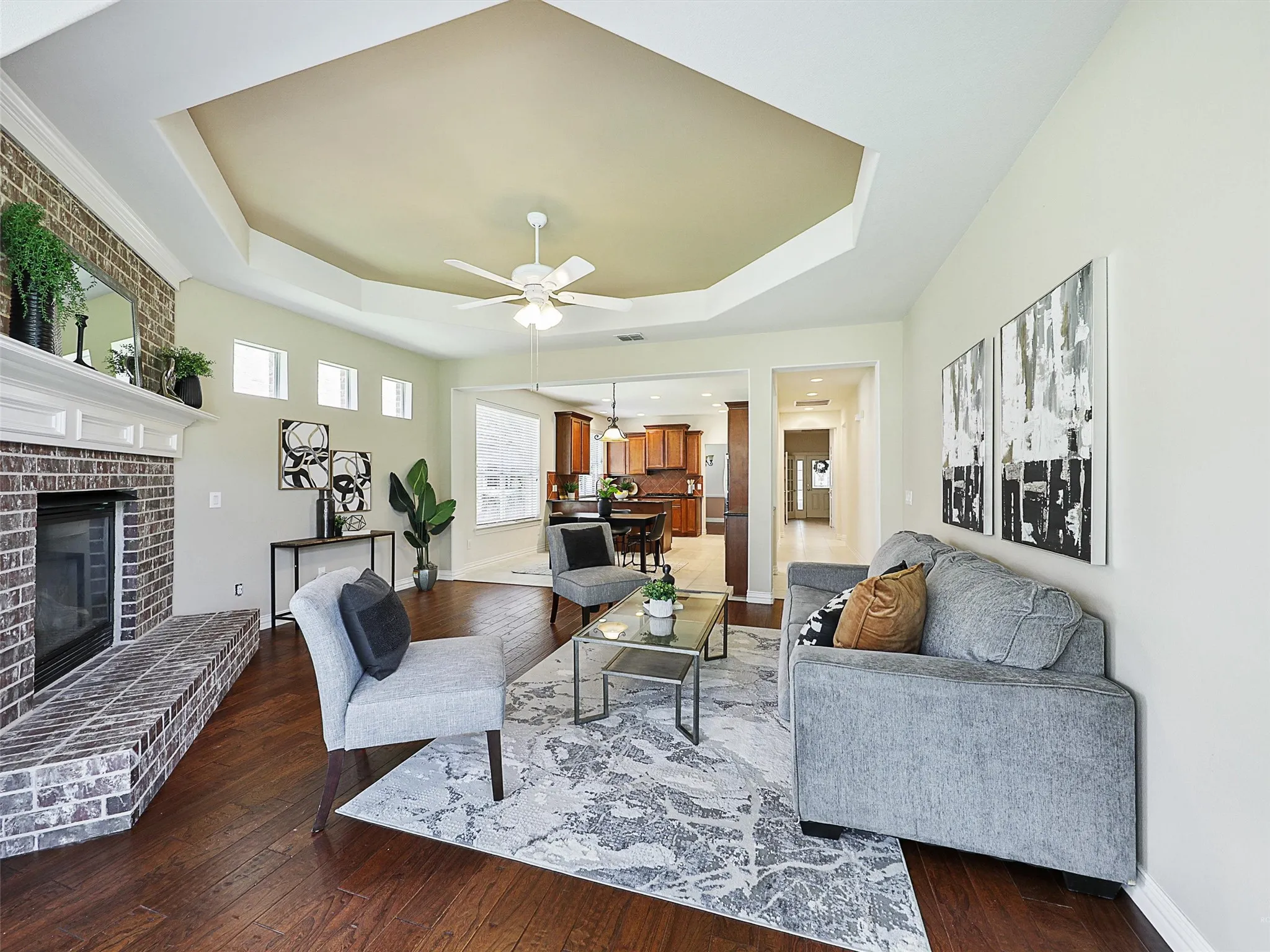 Living area with a raised ceiling, dark wood-style flooring, a brick fireplace, and ceiling fan