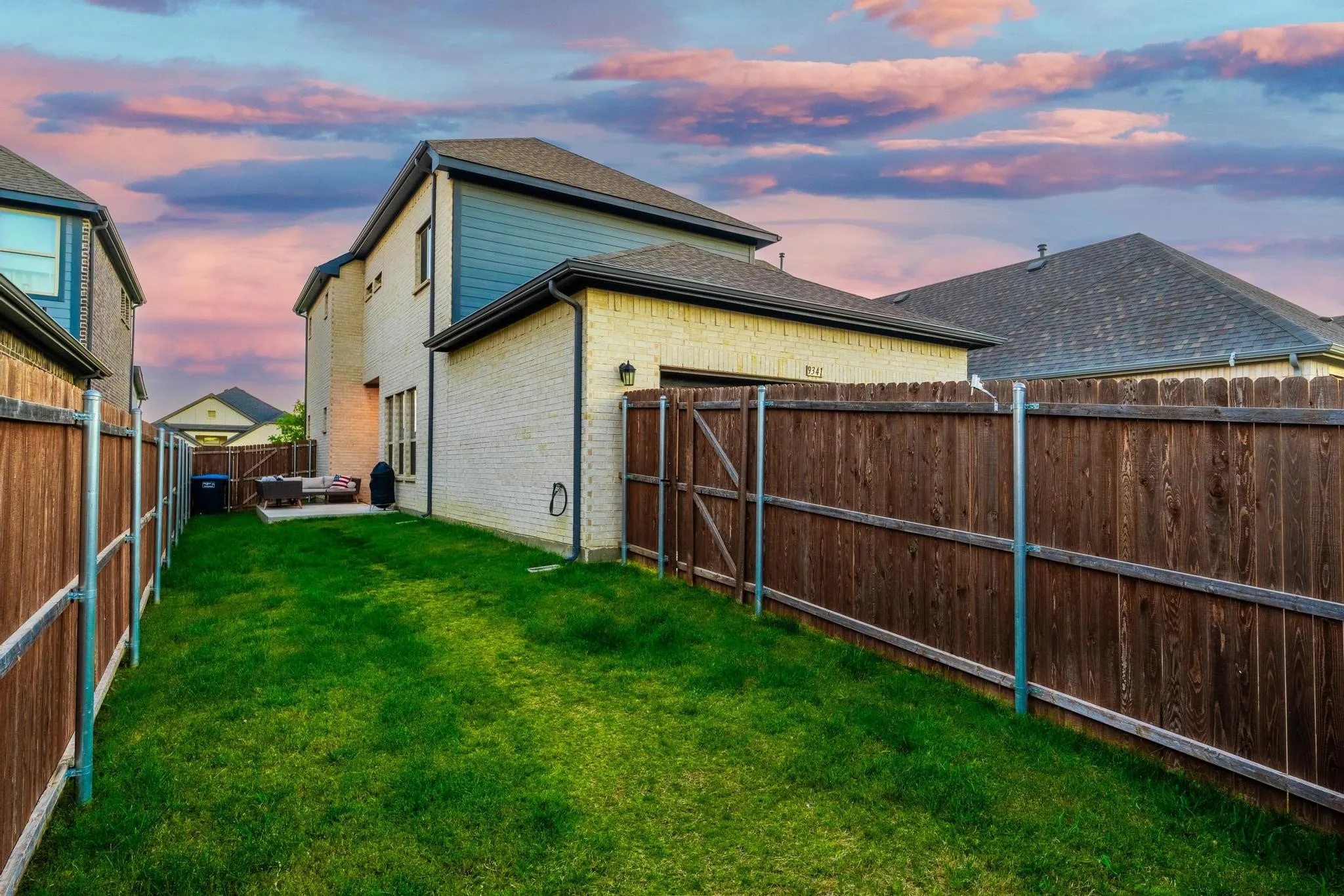 Grassed backyard area featuring a concrete patio, brick exterior, dark siding accents, and a privacy fence with gate