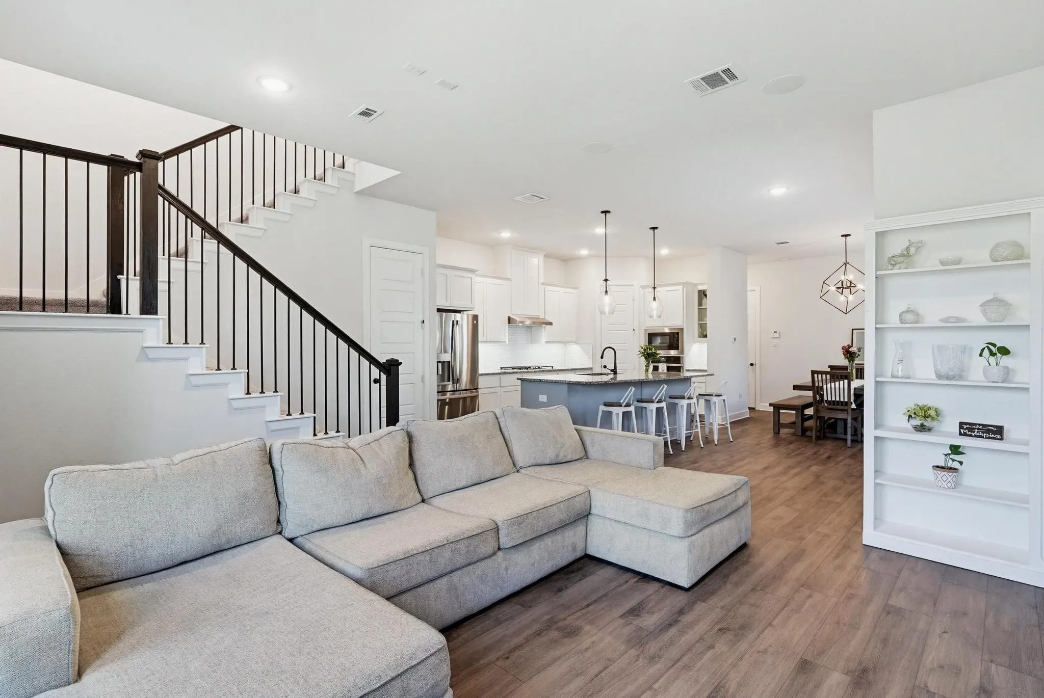 Open-concept living area featuring wood-finish flooring, a built-in bookshelf, staircase with dark wood handrail and black metal spindles, and recessed lighting