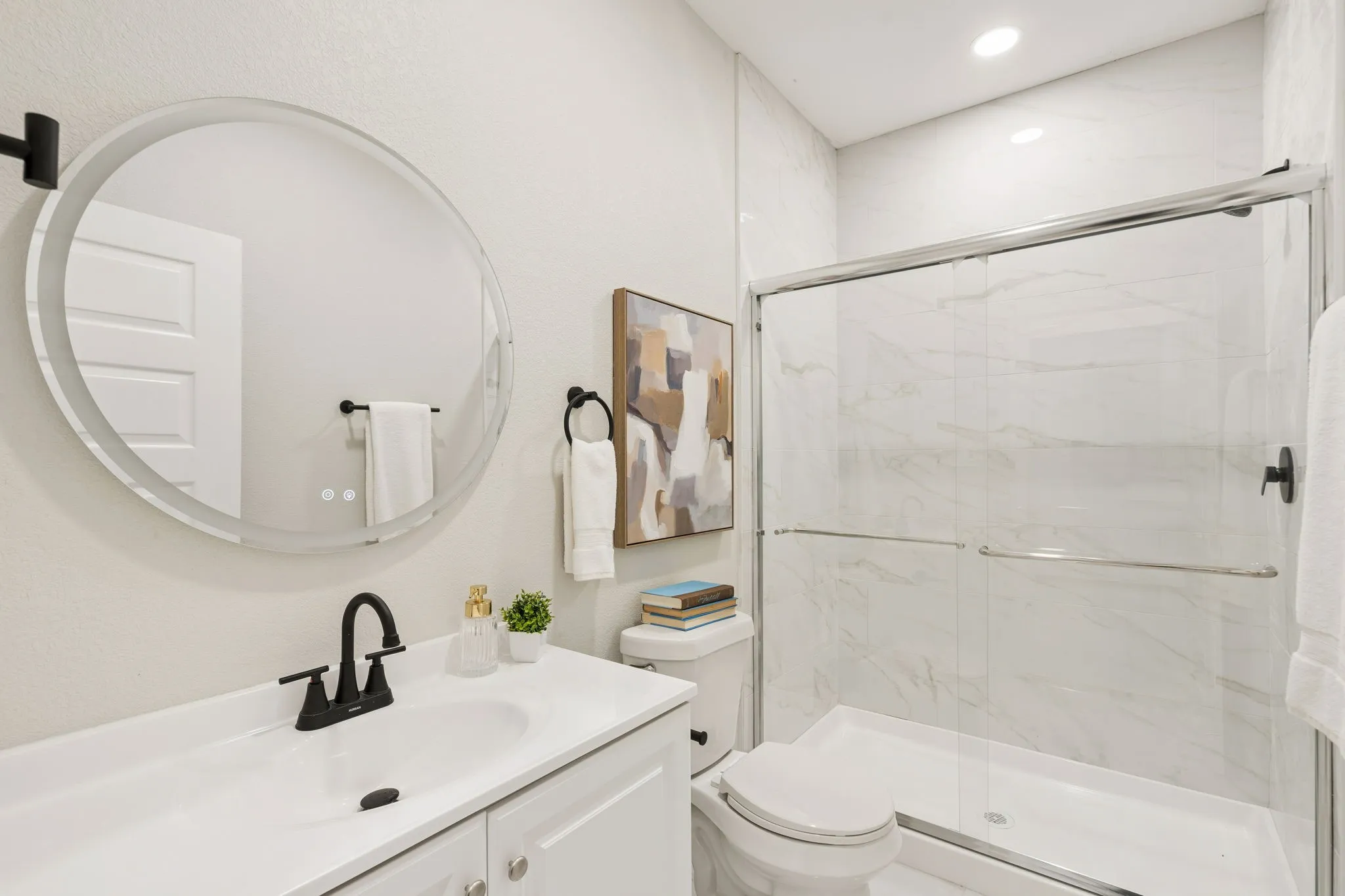 Bathroom featuring a glass-enclosed shower with white tiled walls, a white vanity with integrated sink, a circular illuminated mirror, and matte black fixtures