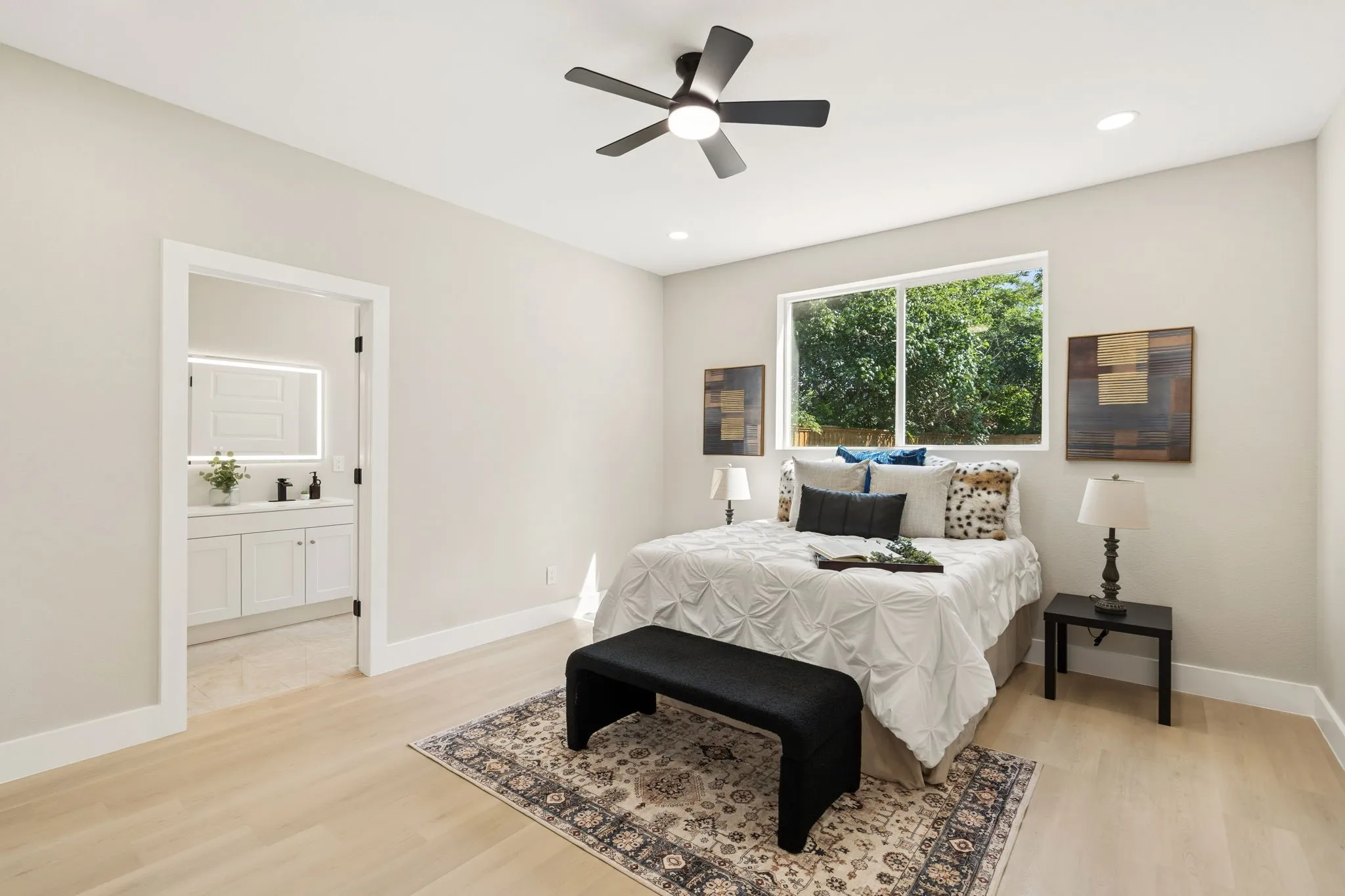 Recessed lighting and a ceiling fan with integrated lighting illuminate this bedroom, featuring light wood-finish flooring and a bright window with exterior tree foliage