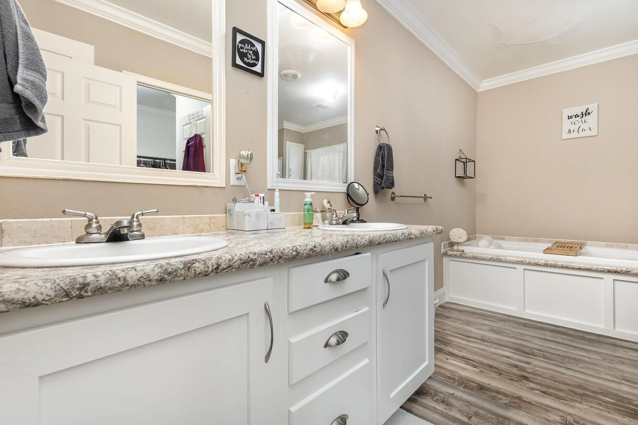 Bathroom featuring a dual vanity with white cabinetry and speckled countertops, a built-in tub with white paneling, wood-finish flooring, and crown molding