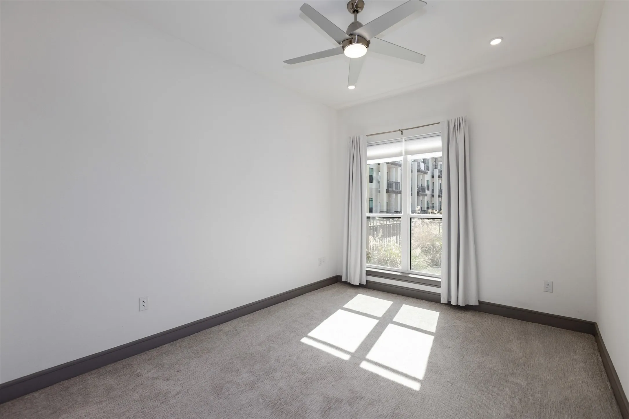 Light-filled room featuring a large window with roller shades and curtains, a ceiling fan with integrated lighting, recessed lighting, and grey carpeting