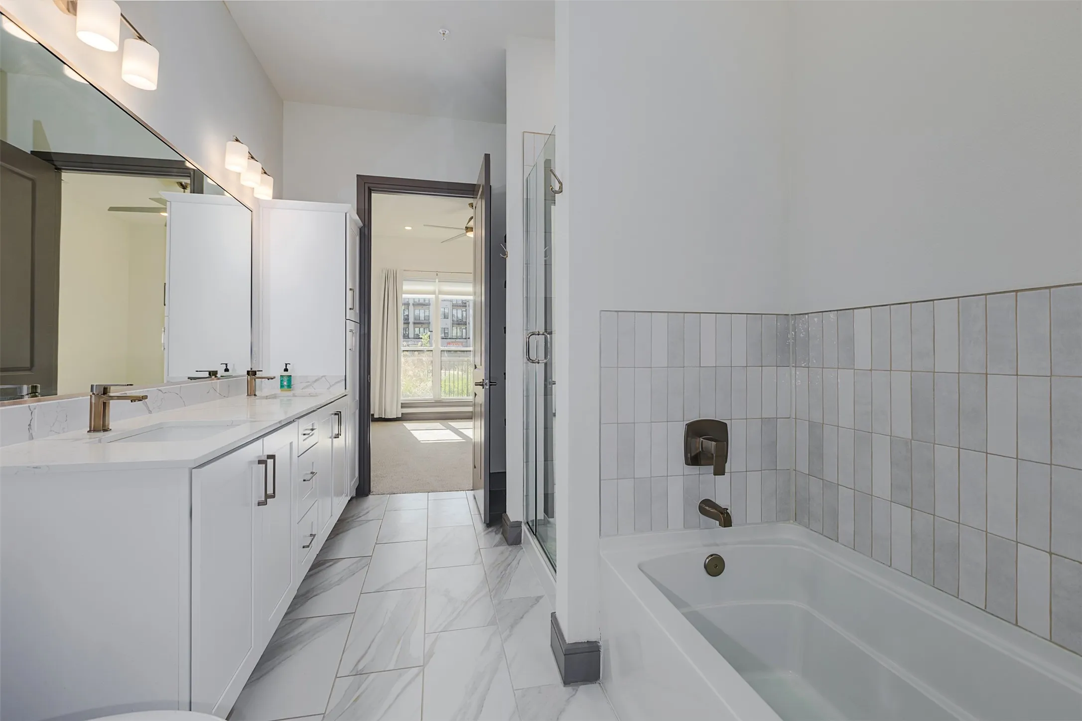 Bathroom featuring a double vanity with white cabinetry, a built-in bathtub with vertical tile surround, a glass-enclosed shower, and large format floor tiling