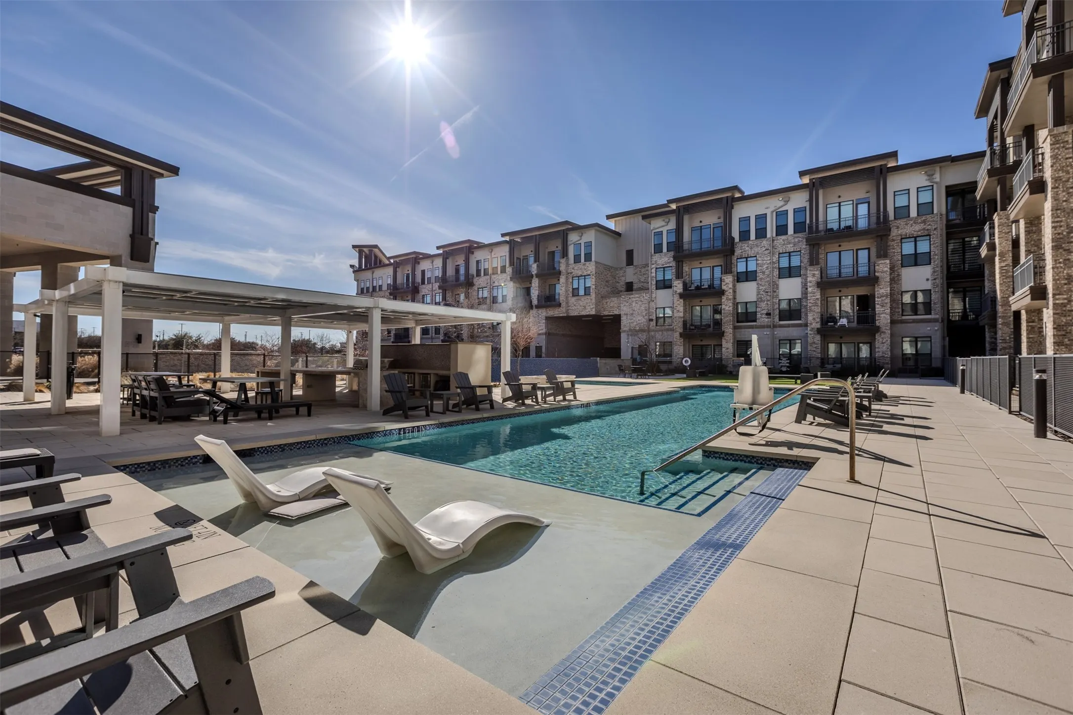 Resort-style swimming pool featuring in-pool lounge seating, a shaded pergola with outdoor kitchen, surrounding sun deck, and multi-story residential buildings with private balconies