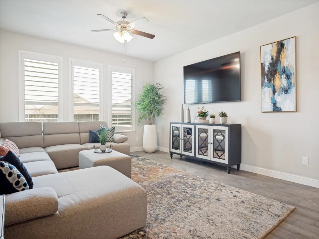 Living area featuring wood-finish flooring, a ceiling fan with integrated lighting, and multiple windows fitted with louvered shutters