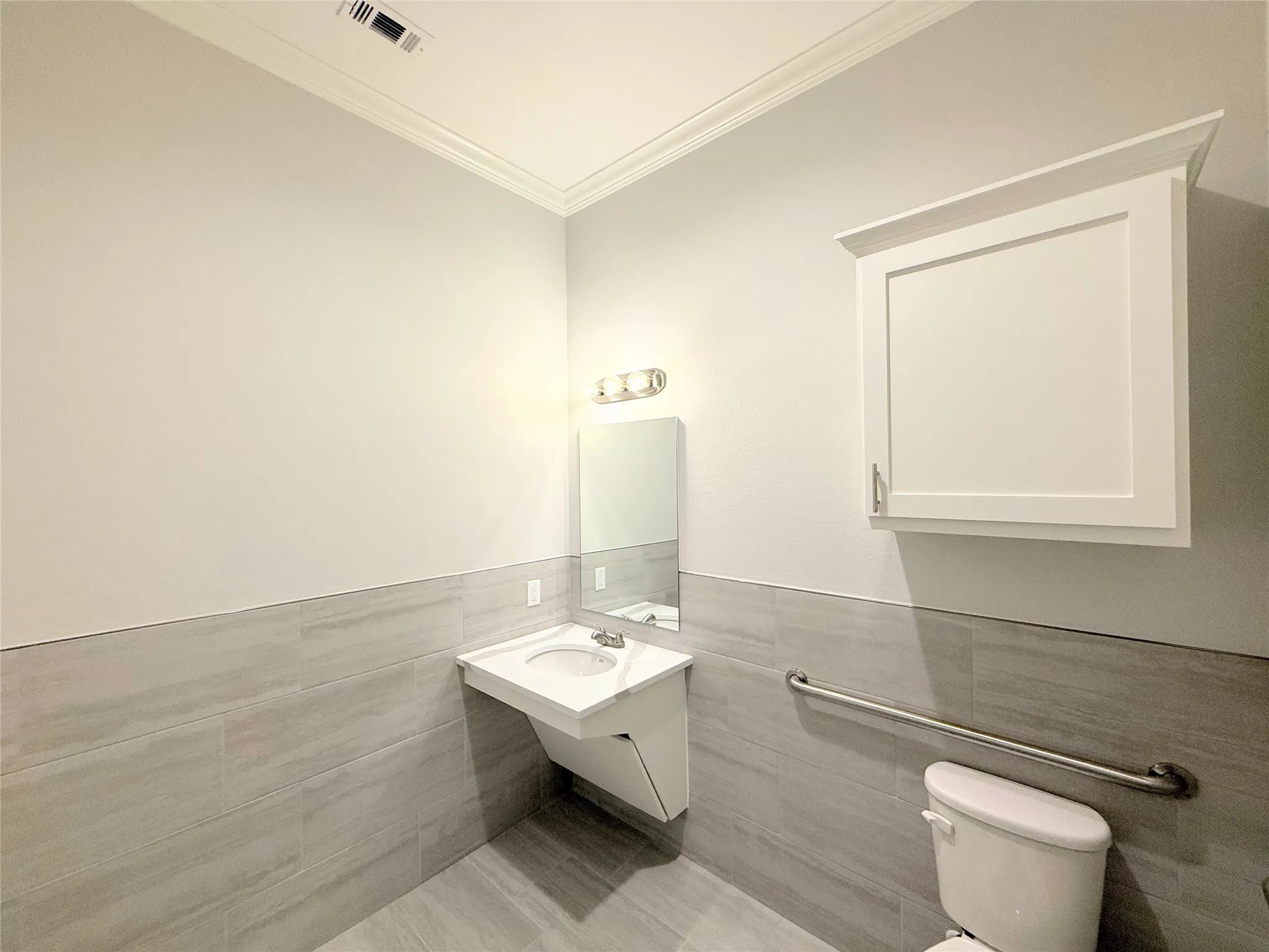 Bathroom featuring light gray wall tile, a floating vanity with integrated sink, a rectangular wall mirror, and a white wall cabinet with crown molding