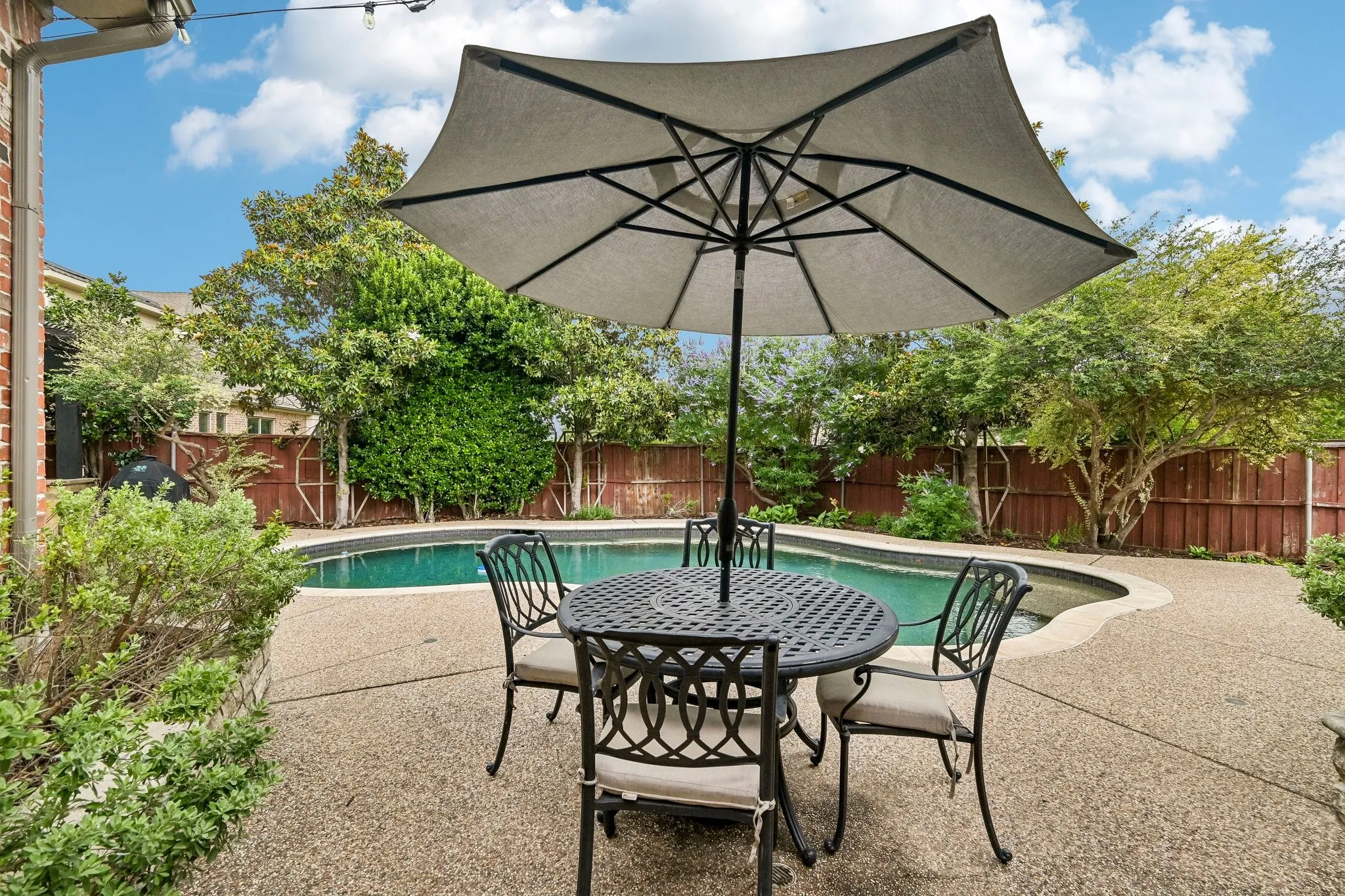 Curved swimming pool featuring a textured concrete surround, mature perimeter landscaping, and a red brick exterior