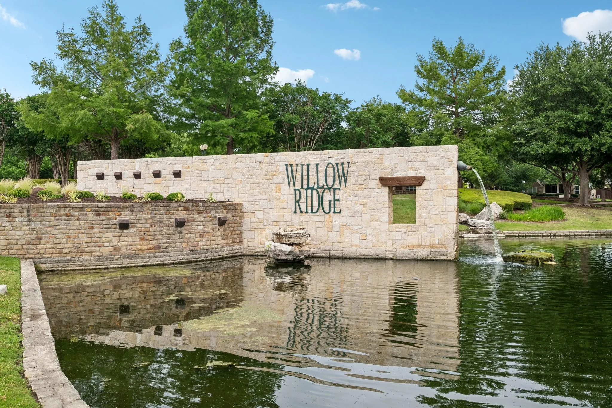 Stone community sign featuring a water feature with a fountain, surrounded by mature trees and lush landscaping
