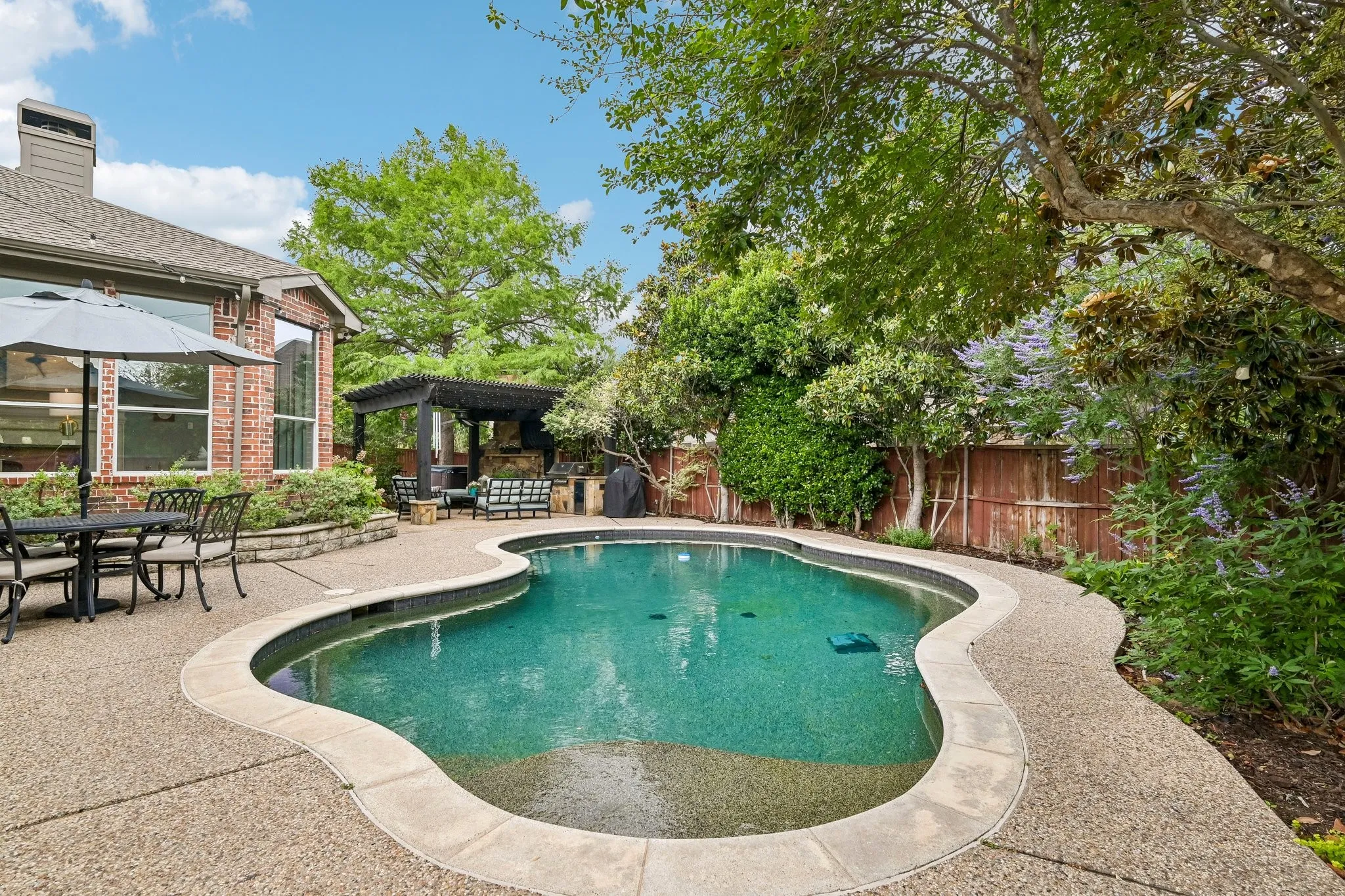 Freeform swimming pool with exposed aggregate concrete decking, surrounded by mature landscaping and a privacy fence