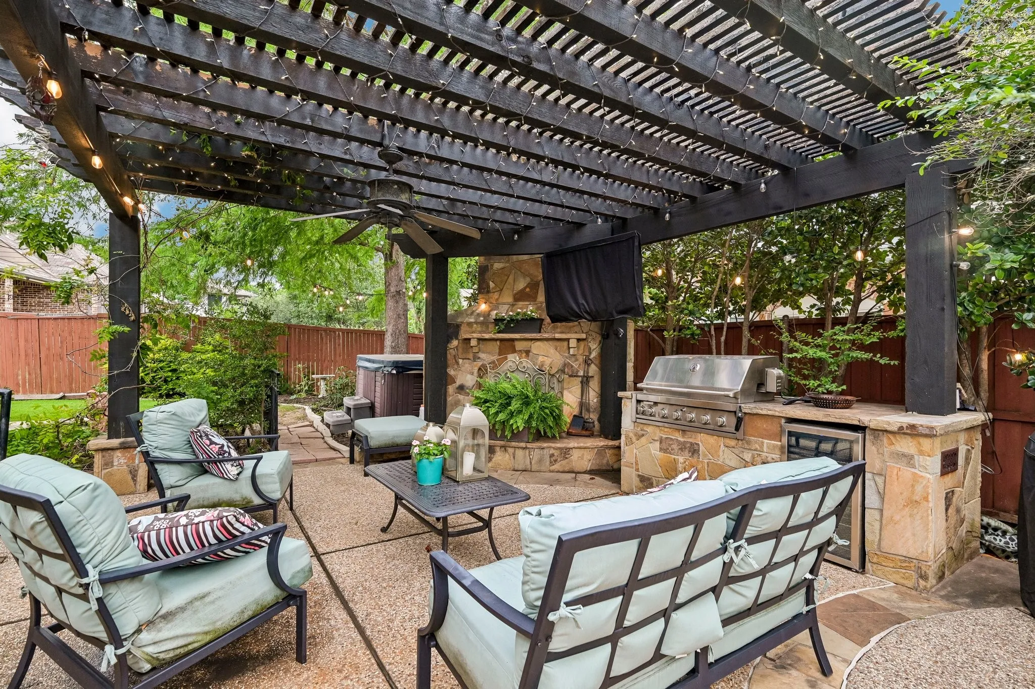 Expansive outdoor living area featuring a dark-stained wood pergola, integrated outdoor kitchen with built-in grill and refrigerator, stone-clad fireplace with mounted outdoor television, ceiling fan, and a built-in spa