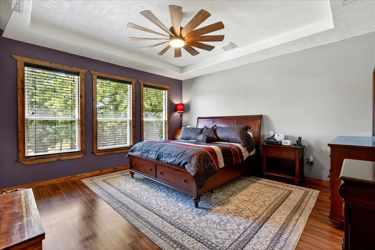 Primary bedroom featuring wood-finish flooring, a tray ceiling, and a farm style ceiling fan with integrated lighting, pretty outdoor view from the windows.
