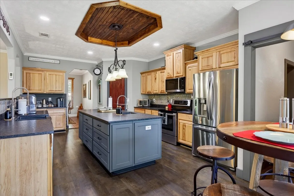 Spacious kitchen featuring a central island with integrated storage, wood-finish flooring, recessed lighting, and a wood-paneled ceiling accent. Open to the living, granite counters, pine cabinets, tile back splash, double black metal farm sink, coffee bar with small sink, large pantry and breakfast area.