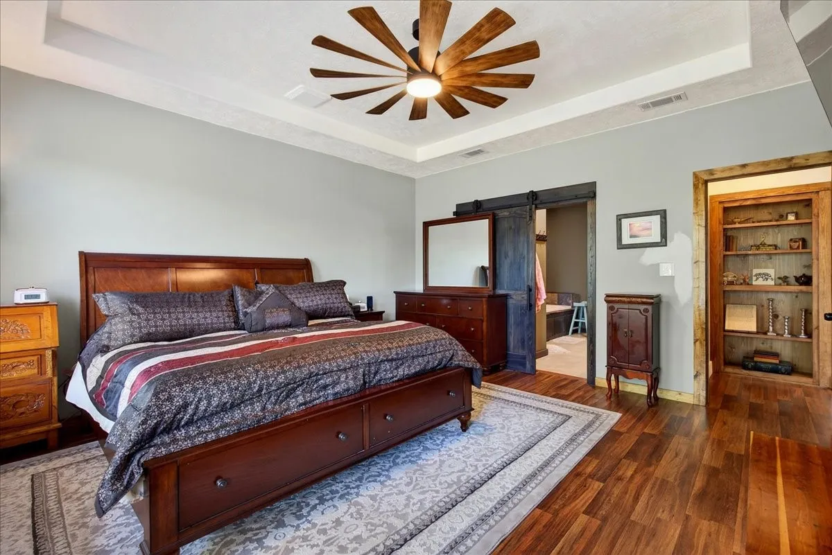 Primary bedroom featuring wood-finish flooring, a tray ceiling, and a farm style ceiling fan with integrated lighting, pretty outdoor view from the windows.