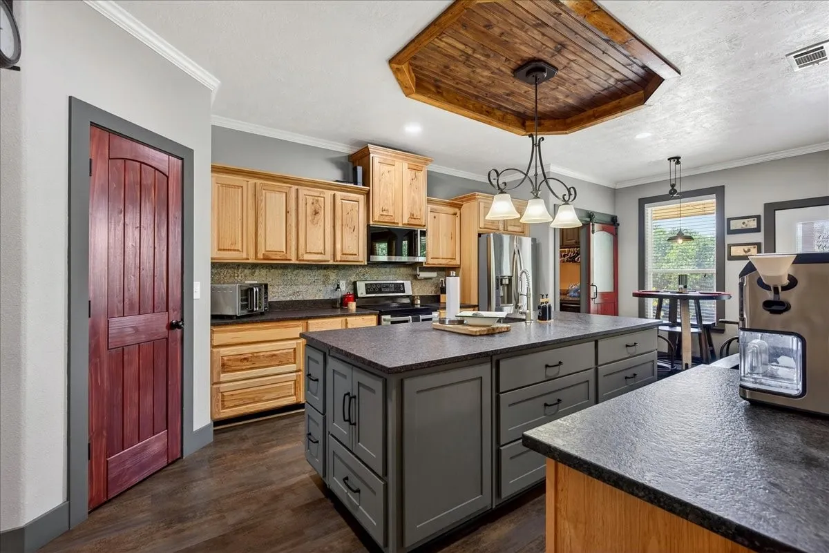 Kitchen featuring a central island with integrated storage, wood-finish flooring, light wood cabinetry, stone-look countertops, and a wood-paneled ceiling accents, granite counters, pine cabinets, tile back splash, double black metal farm sink, large pantry.