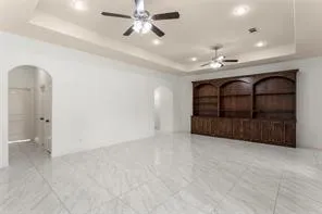 Spacious room featuring white tile flooring, a dark wood built-in wall unit with arched shelving, multiple ceiling fans, recessed lighting, and a tray ceiling
