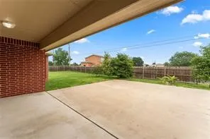Covered concrete patio extending to a grass lawn, featuring a brick exterior wall, wood fencing, and mature landscaping