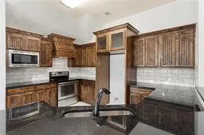 Kitchen featuring rich wood cabinetry, dark countertops, subway tile backsplash, stainless steel appliances, and an undermount sink with a black faucet