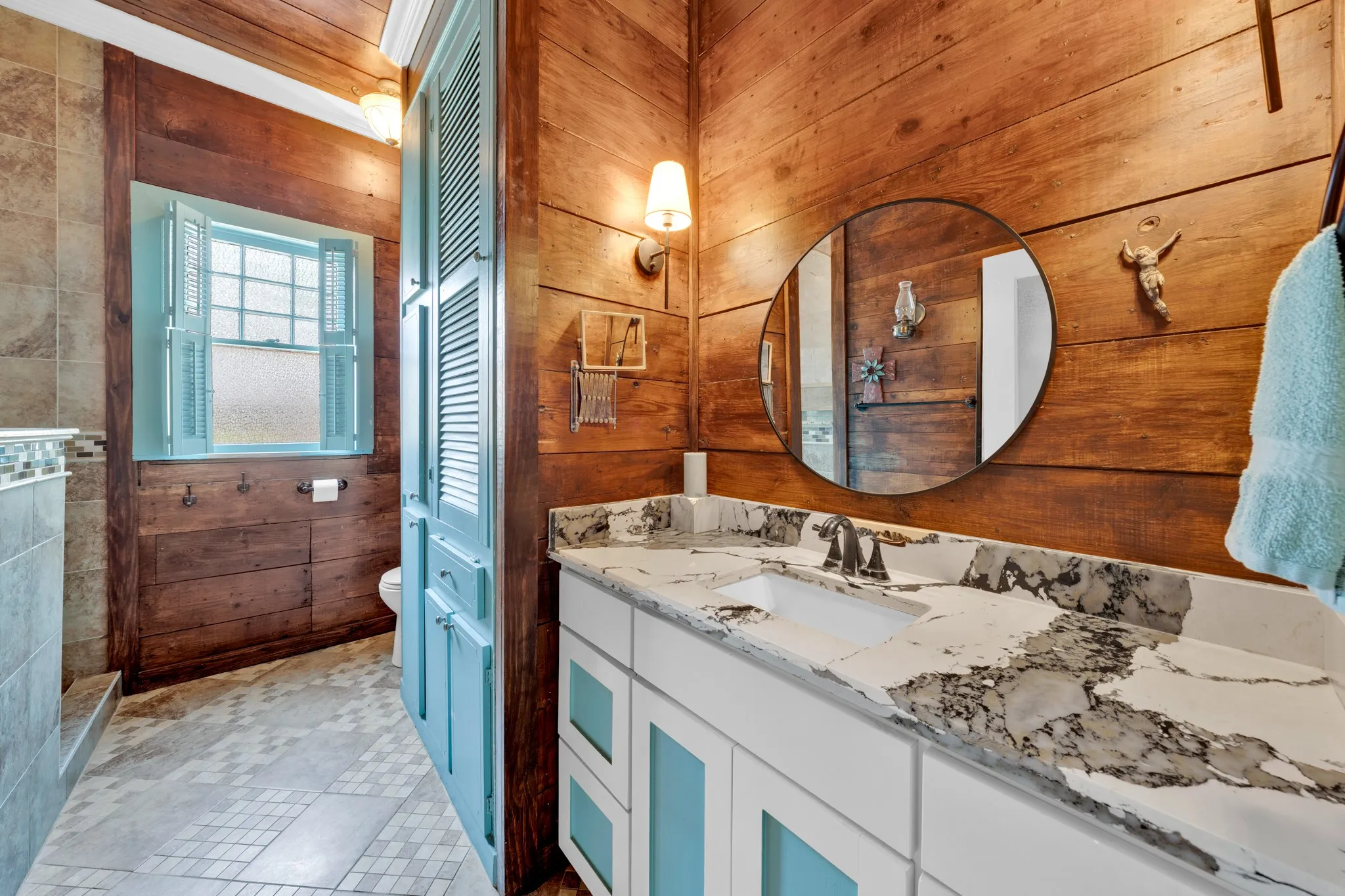 Double vanity with integrated sinks, marble-patterned countertop, wood-finish wall paneling, and a round wall-mounted mirror