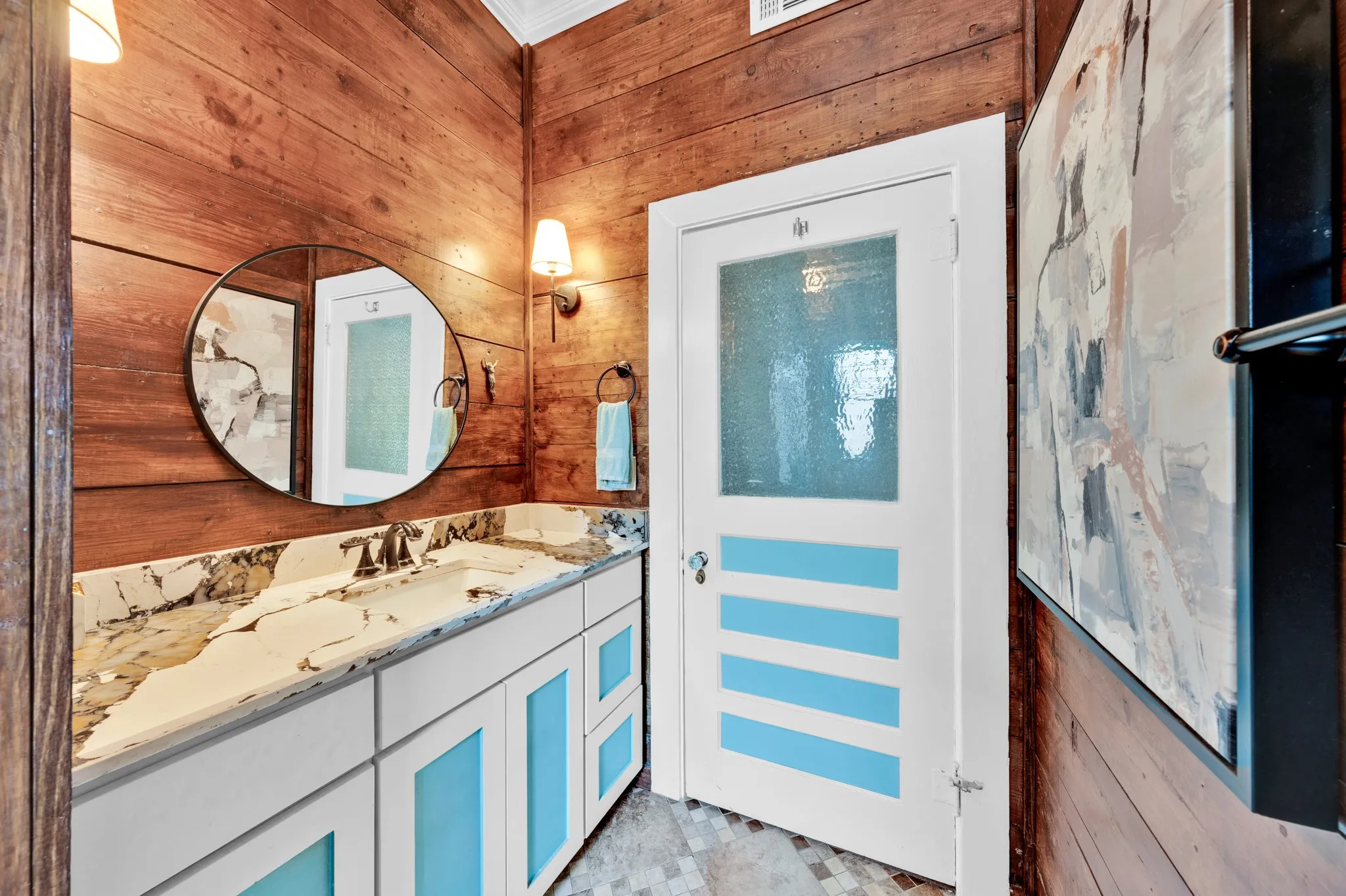 Wood-paneled wall finishes, white vanity with blue-accented doors, built-in sink with stone-finish countertop, round wall mirror, and wall-mounted sconces