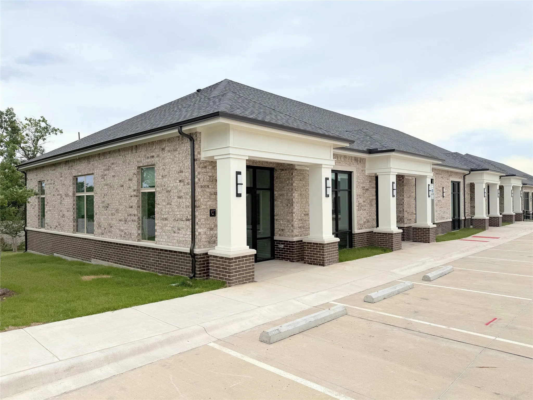 Modern commercial building featuring light-colored brick with dark brick accents, multiple storefront entrances, a dark shingle roof, and a concrete parking area