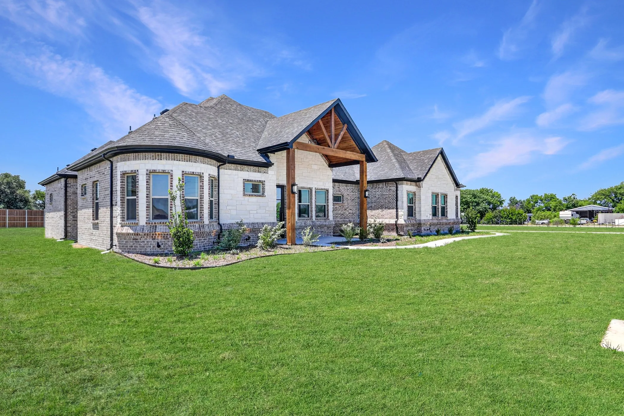 Expansive green lawn bordering a newly constructed home featuring a mixed-material exterior, a gabled roof with dark shingles, and a covered patio with exposed timber beams