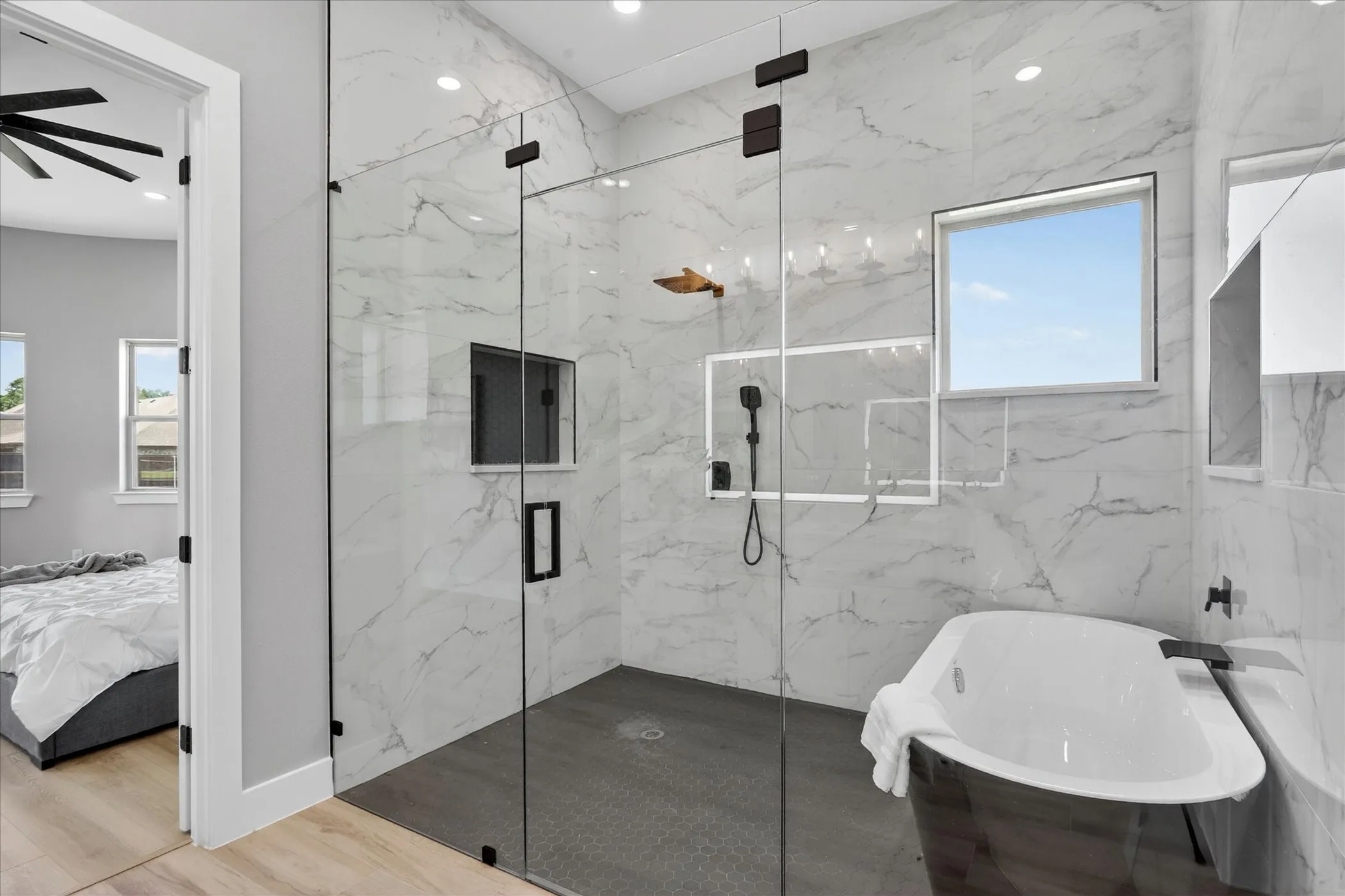 Spacious bathroom featuring a glass-enclosed shower with contrasting dark tile flooring, a freestanding soaking tub, and a window for natural light