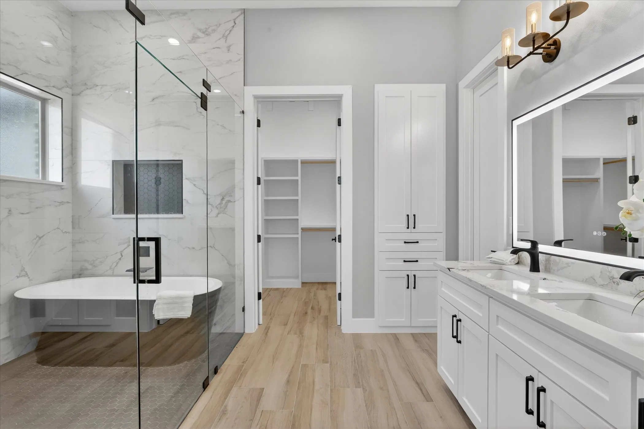 Bathroom featuring a dual vanity with quartz countertop, light wood-finish flooring, a freestanding soaking tub, glass-enclosed shower, and built-in white cabinetry
