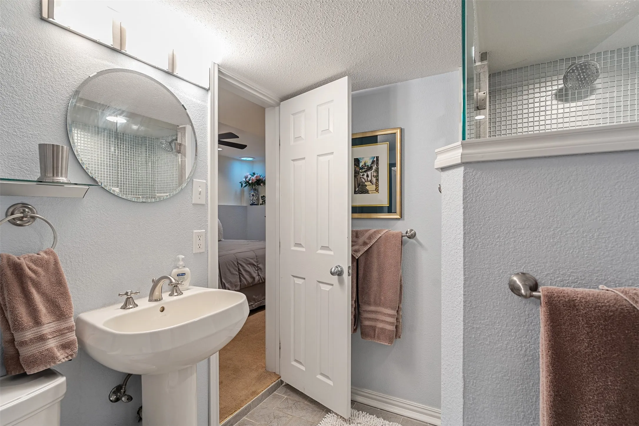 Bathroom featuring a pedestal sink with a brushed nickel faucet, a round wall-mounted mirror, and a glass-enclosed shower with small square tile