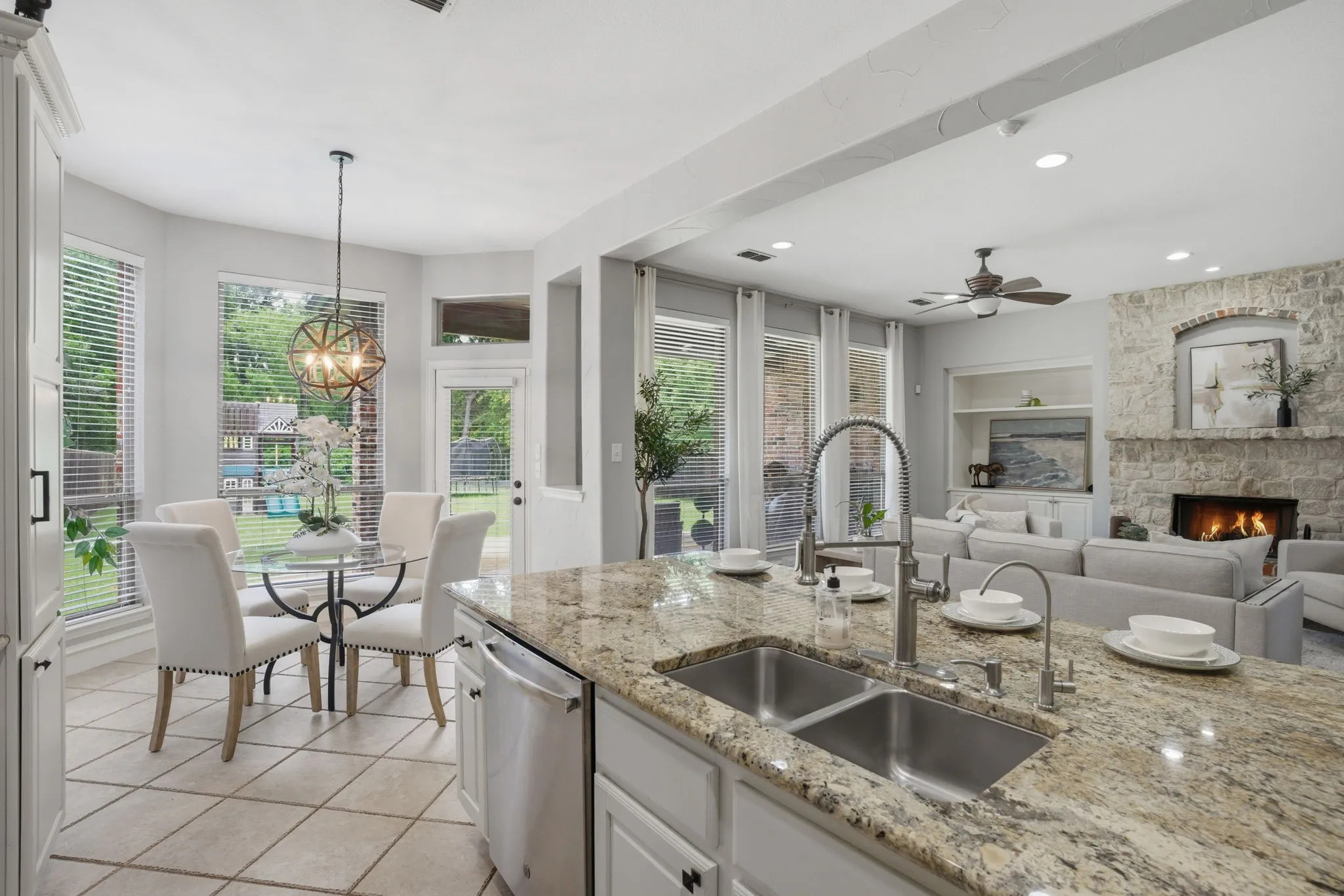Open-concept kitchen and living area featuring a stone fireplace, granite countertops, a double basin stainless steel sink, tile flooring, and abundant natural light from multiple windows