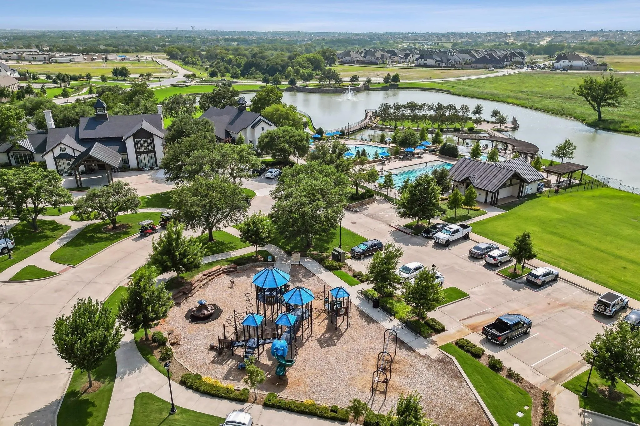 MUSTANG LAKES CLUB HOUSE - Expansive aerial view showcasing a large community lake with a fountain, multiple swimming pools, a central clubhouse, and an outdoor playground