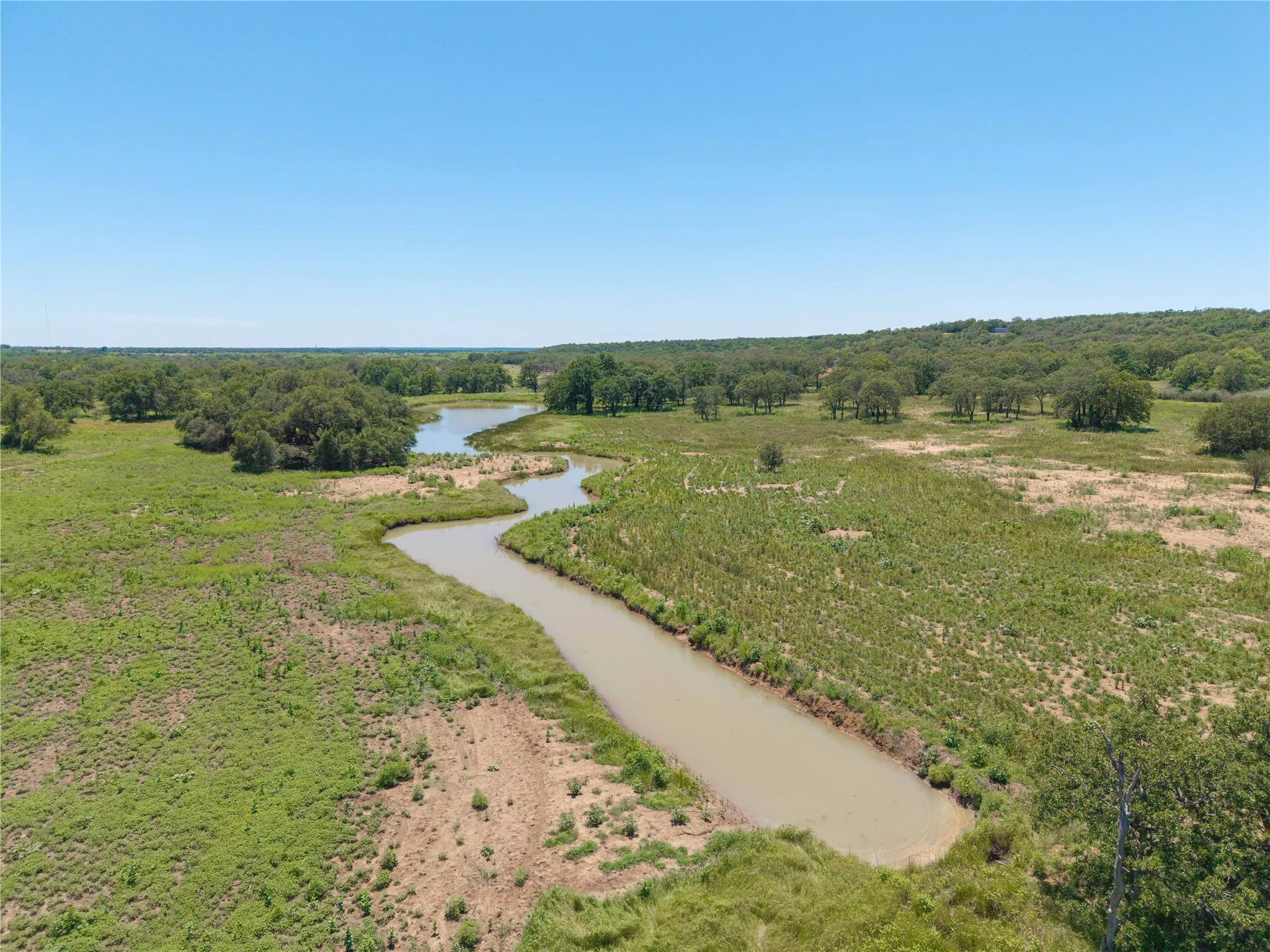 Expansive natural landscape featuring a winding waterway, abundant mature trees, varied green foliage, and open grassland