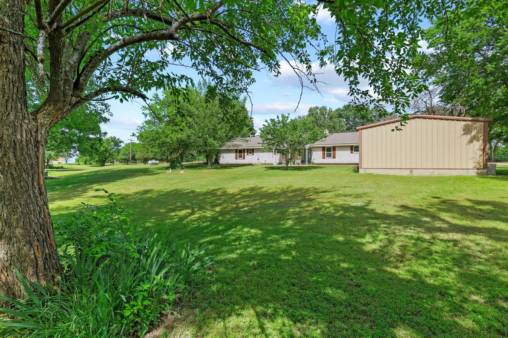 Expansive green lawn with mature trees, featuring a detached metal-sided outbuilding and a single-story residence with a red roof